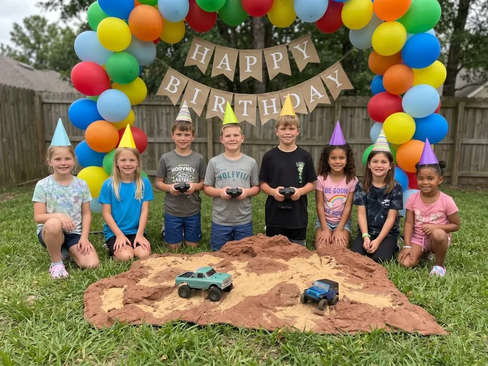 Eight kids in party hats kneeling behind a rock terrain course with two RC trucks under a balloon arch